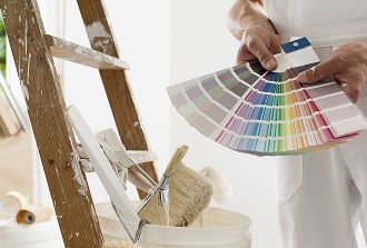 Peintre professionnel tenant un nuancier de couleurs devant des outils de peinture et une échelle en bois tachée.