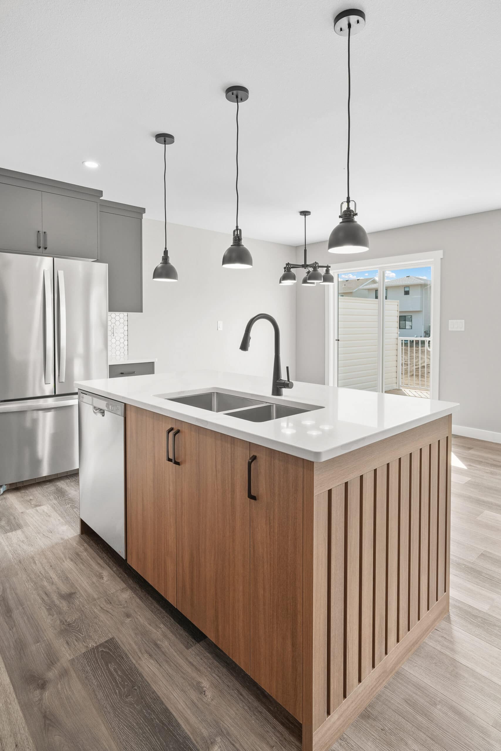 Cuisine moderne avec îlot central bois et comptoir blanc, éclairage suspendu noir, électroménagers acier inoxydable et porte-patio.