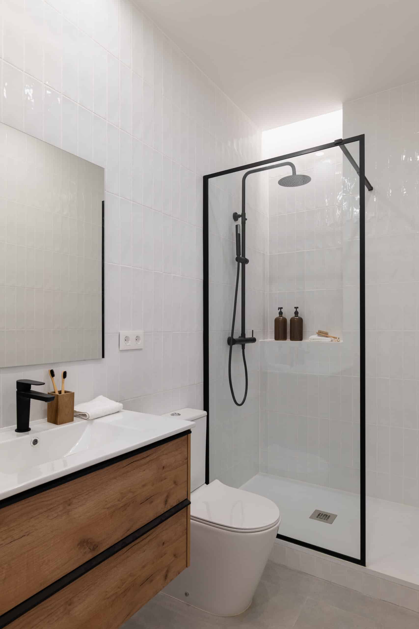 Salle de bain moderne avec carrelage blanc vertical, meuble-lavabo bois, robinetterie noire et douche à l'italienne. Prises murales visibles.
