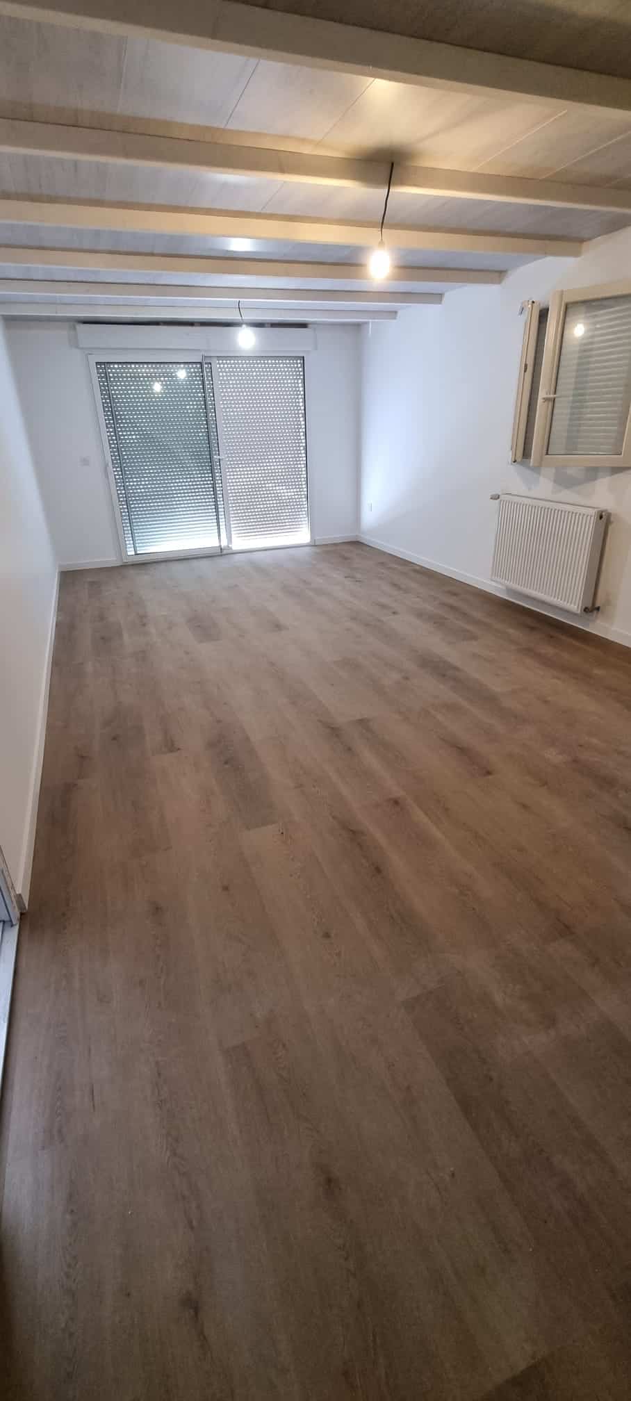 Pièce vide avec parquet flottant brun clair, murs blancs, plafond à poutres apparentes claires et lumière suspendue. Grandes baies vitrées avec volets roulants fermés.