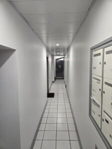 Couloir blanc d'immeuble avec sol carrelé, boîtes aux lettres à droite et porte sombre au fond.