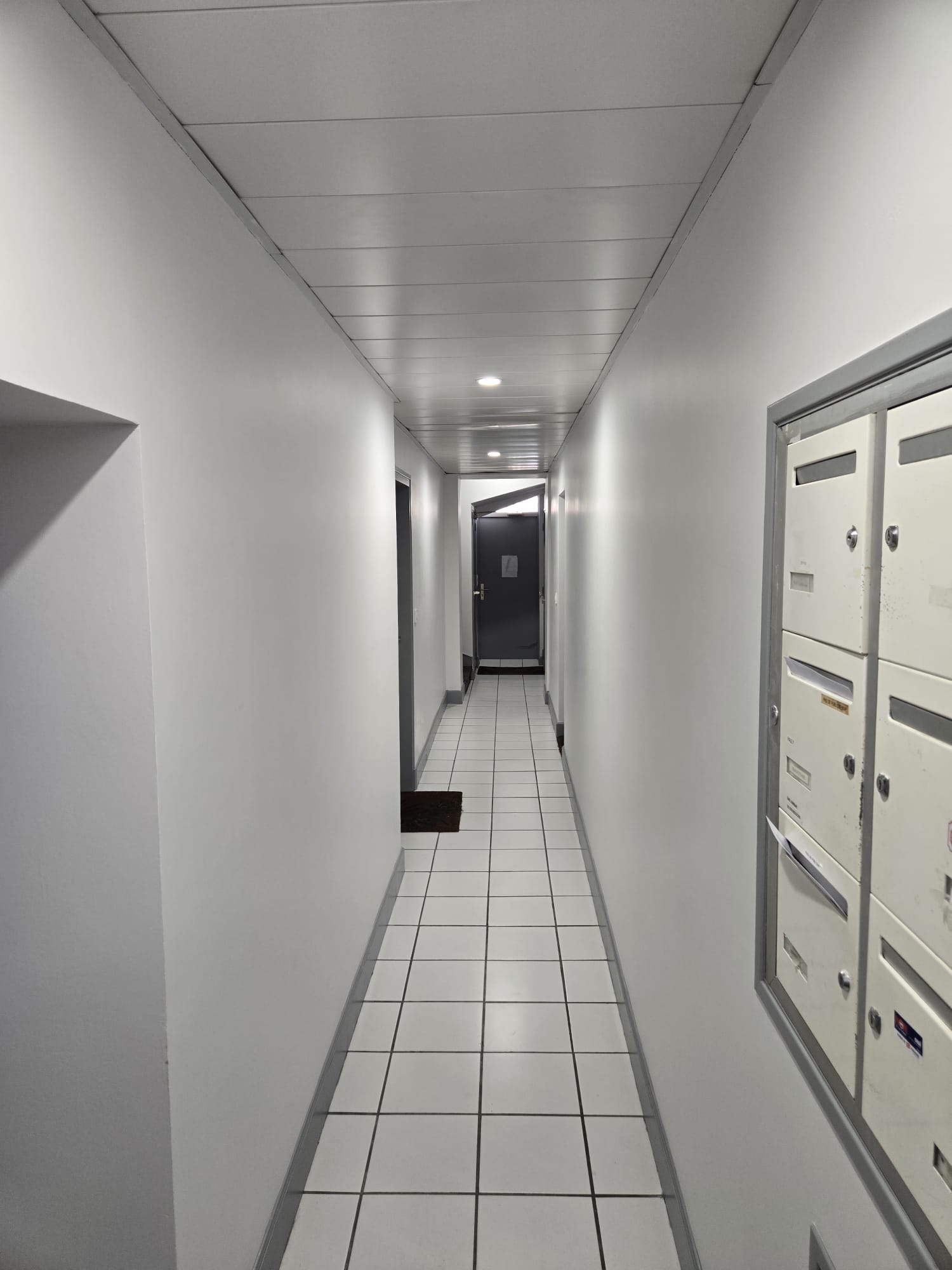 Couloir blanc d'immeuble avec sol carrelé, boîtes aux lettres à droite et porte sombre au fond.