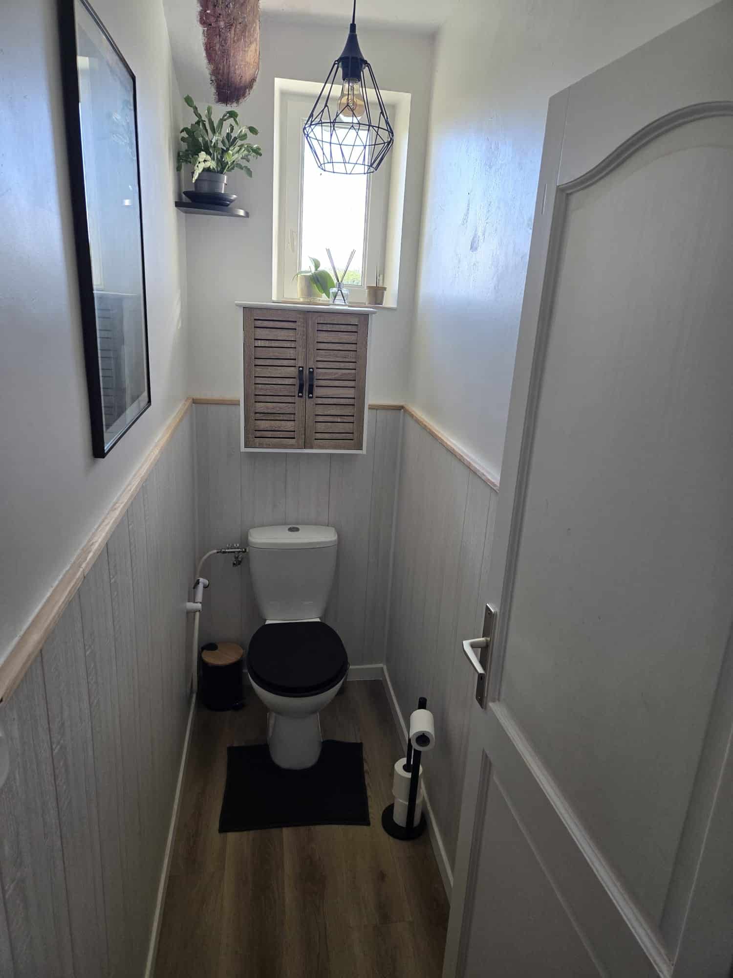 Toilette moderne avec lambris gris, siège noir, étagère en bois et lampe cage industrielle dans un petit espace.