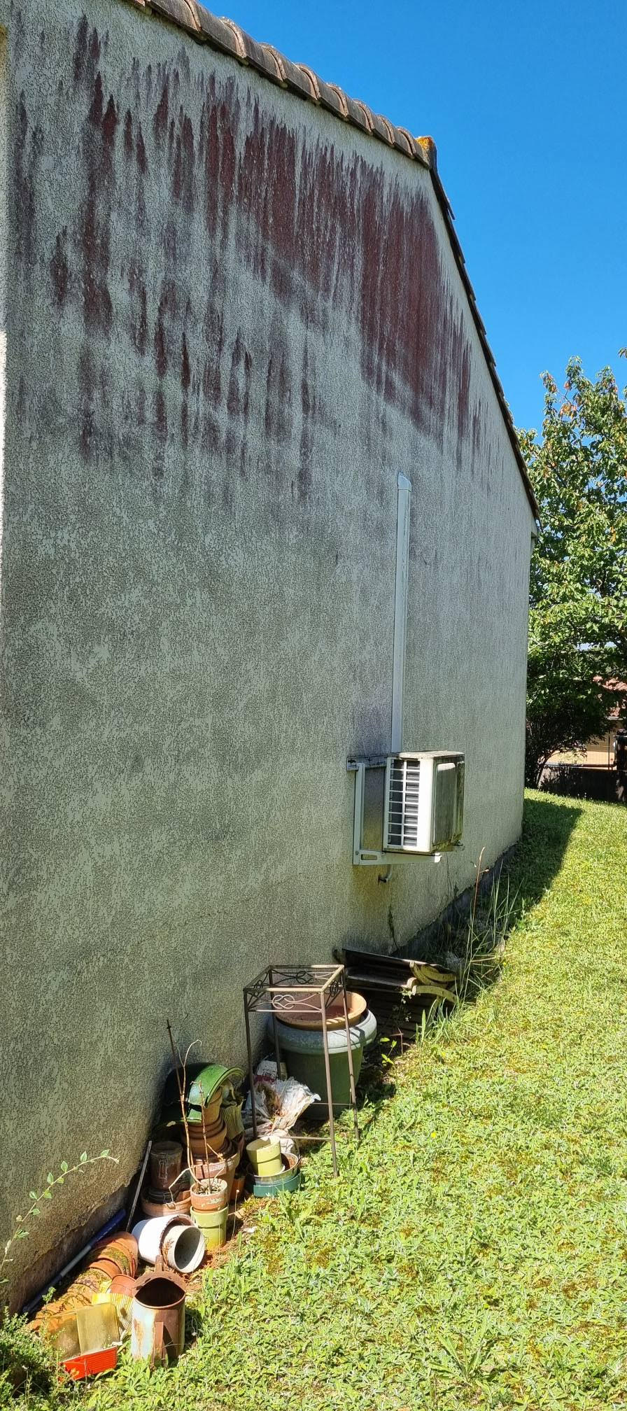 Façade de maison avec d'importantes traces d'algues rouges et noires, climatiseur mural et pots de fleurs au sol.