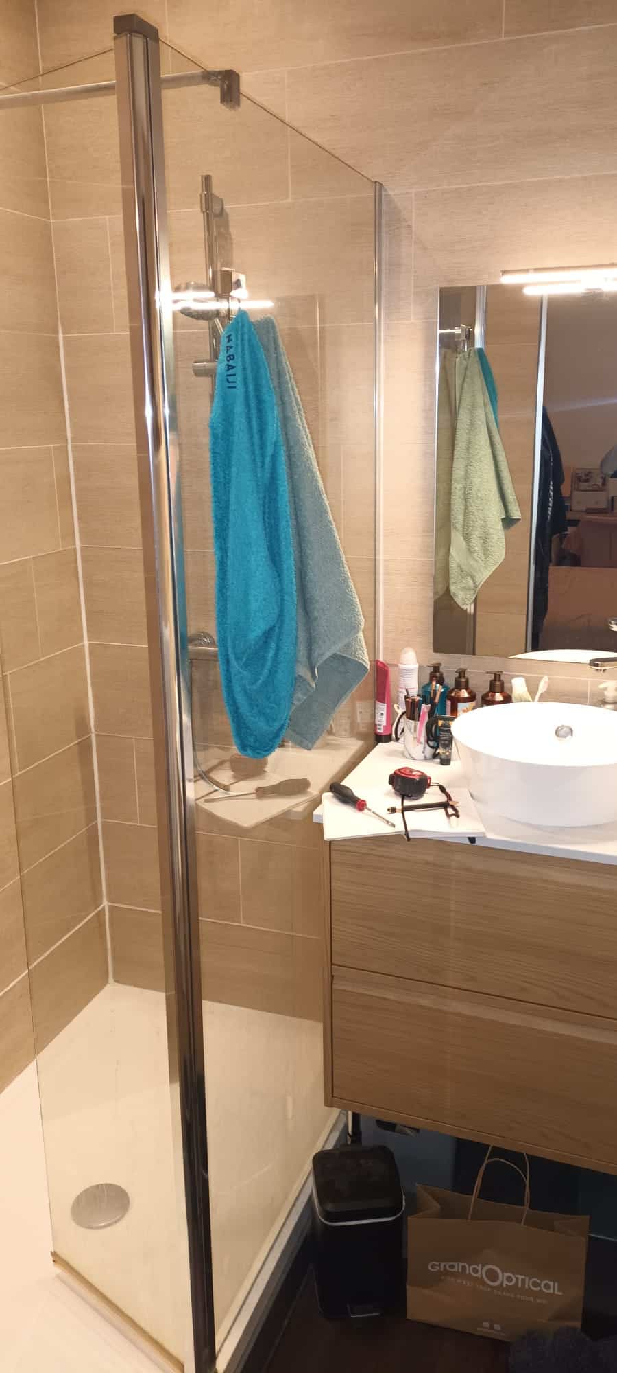 Douche italienne, paroi verre, meuble bois, serviette Nabaiji et sac GrandOptical dans une salle de bain moderne.